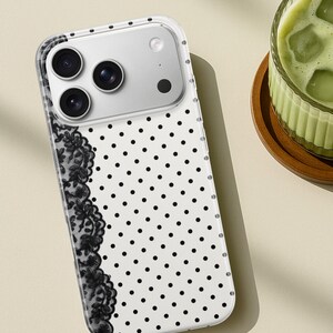 May include: Clear phone case with a white background and black polka dots. A black lace design is on the left side. The phone case has a camera module with three lenses and a flash.