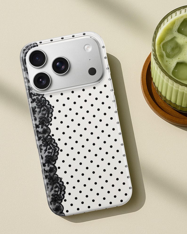 May include: Clear phone case with a white background and black polka dots. A black lace design is on the left side. The phone case has a camera module with three lenses and a flash.