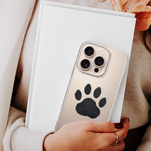 May include: A clear phone case with a black paw print design. The phone case is held in front of a white book. The phone has three camera lenses. The person holding the phone is wearing a cream-colored sweater and blazer.