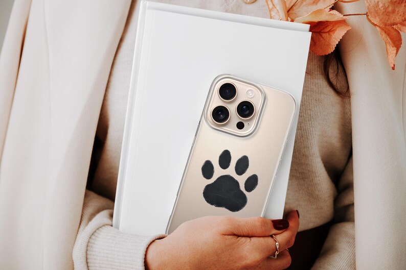 May include: A clear phone case with a black paw print design. The phone case is held in front of a white book. The phone has three camera lenses. The person holding the phone is wearing a cream-colored sweater and blazer.