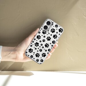 May include: Clear phone case featuring a pattern of black paw prints on a white background. The case is held in a hand, showcasing the design. The case is designed to fit a smartphone.