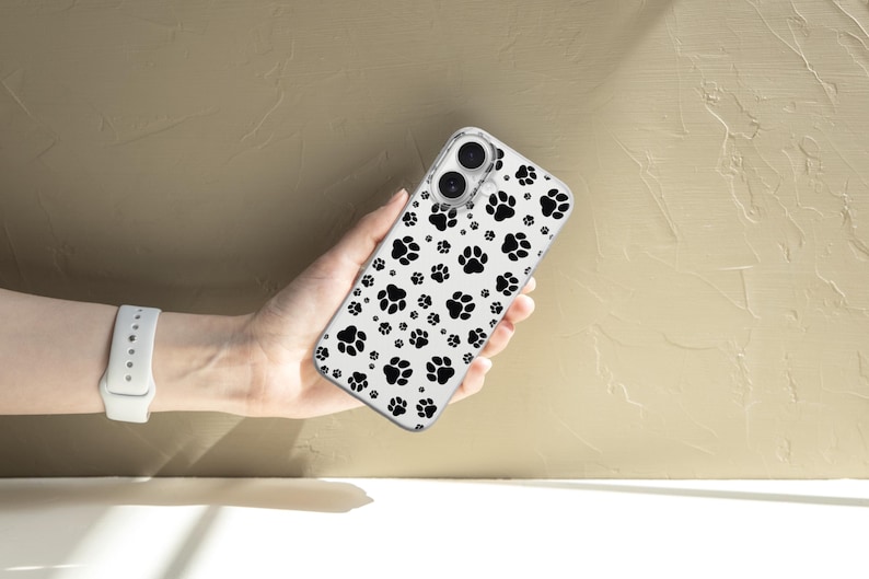 May include: Clear phone case featuring a pattern of black paw prints on a white background. The case is held in a hand, showcasing the design. The case is designed to fit a smartphone.
