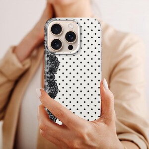 May include: A white iPhone case with a black polka dot pattern and a black lace design on the left side. The phone case is held by a person wearing a tan blazer.