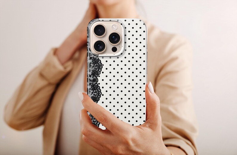 May include: A white iPhone case with a black polka dot pattern and a black lace design on the left side. The phone case is held by a person wearing a tan blazer.