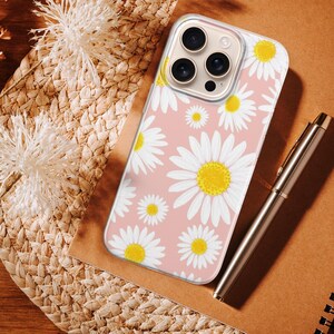 Pode incluir: Uma capa de telefone com um padr&atilde;o de margaridas brancas e amarelas sobre um fundo rosa claro. Uma caneta prateada e um caderno espiral est&atilde;o ao lado da capa. A capa est&aacute; sobre uma esteira de palha tecida.