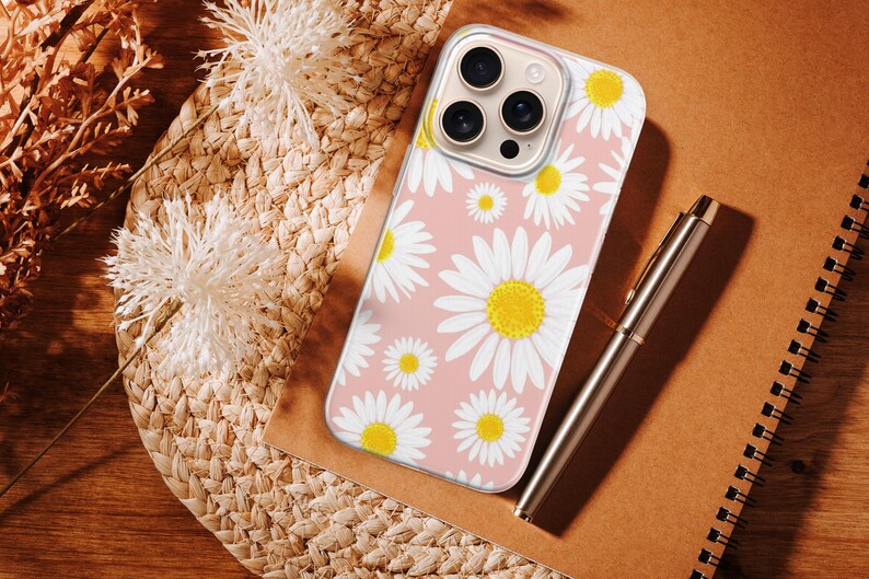 Pode incluir: Uma capa de telefone com um padr&atilde;o de margaridas brancas e amarelas sobre um fundo rosa claro. Uma caneta prateada e um caderno espiral est&atilde;o ao lado da capa. A capa est&aacute; sobre uma esteira de palha tecida.