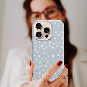 Pode incluir: Capa de telefone azul claro com um padr&atilde;o de margaridas. A capa tem uma borda transparente e um recorte para a c&acirc;mera. As margaridas s&atilde;o brancas com centros amarelos. A capa do telefone &eacute; segurada em uma m&atilde;o com esmalte de unhas vermelho.