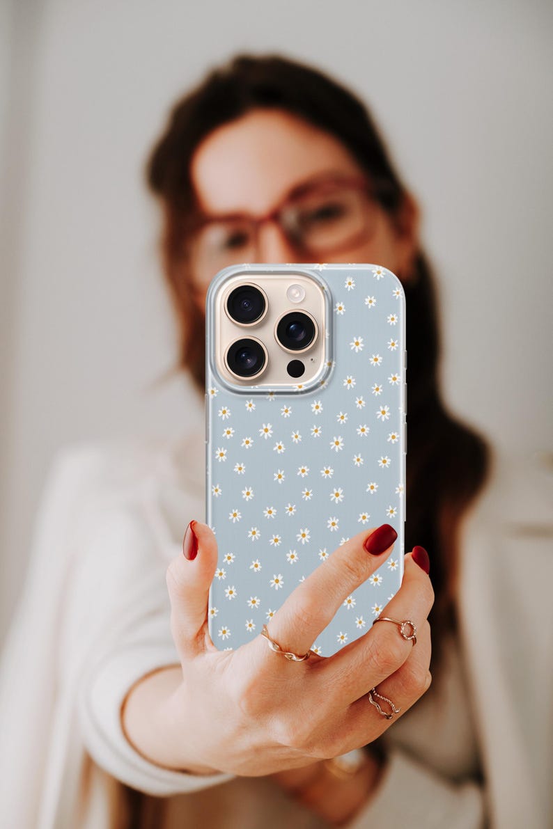 Pode incluir: Capa de telefone azul claro com um padr&atilde;o de margaridas. A capa tem uma borda transparente e um recorte para a c&acirc;mera. As margaridas s&atilde;o brancas com centros amarelos. A capa do telefone &eacute; segurada em uma m&atilde;o com esmalte de unhas vermelho.