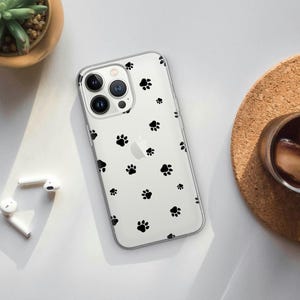 May include: Clear phone case with a pattern of black paw prints. The case is on a white surface with a small plant, a cork coaster, and a pair of white wireless earbuds.