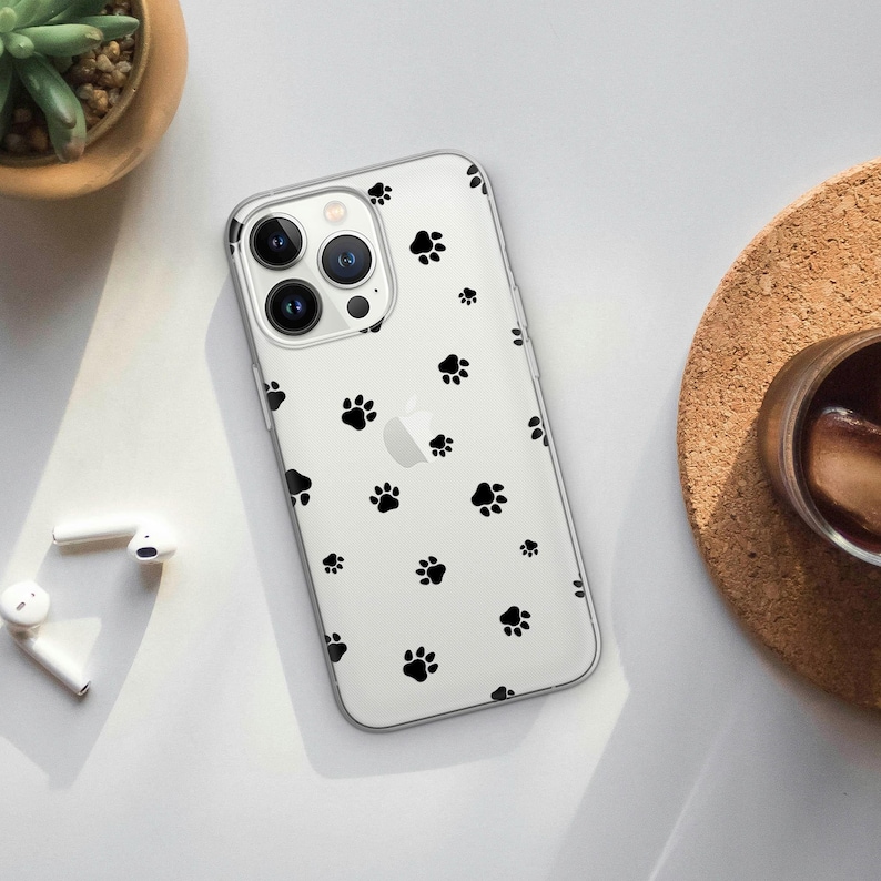 May include: Clear phone case with a pattern of black paw prints. The case is on a white surface with a small plant, a cork coaster, and a pair of white wireless earbuds.