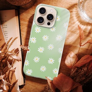 Pode incluir: Uma capa de telefone verde claro com um padr&atilde;o de margaridas. A capa est&aacute; sobre uma superf&iacute;cie de madeira com flores secas, um livro e um copo de &aacute;gua. As margaridas s&atilde;o brancas com centros amarelos.