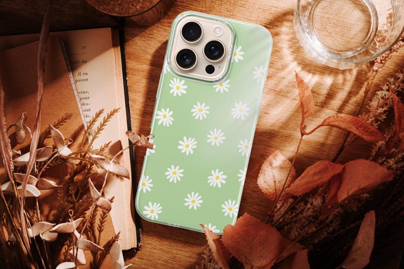 Pode incluir: Uma capa de telefone verde claro com um padr&atilde;o de margaridas. A capa est&aacute; sobre uma superf&iacute;cie de madeira com flores secas, um livro e um copo de &aacute;gua. As margaridas s&atilde;o brancas com centros amarelos.