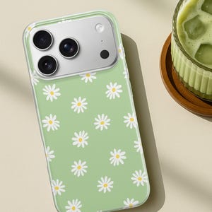 Pode incluir: Uma capa de telefone verde claro com um padr&atilde;o de margaridas. A capa tem uma borda transparente e um recorte para a c&acirc;mera. As margaridas s&atilde;o brancas com centros amarelos. A capa est&aacute; sobre uma superf&iacute;cie marrom claro.