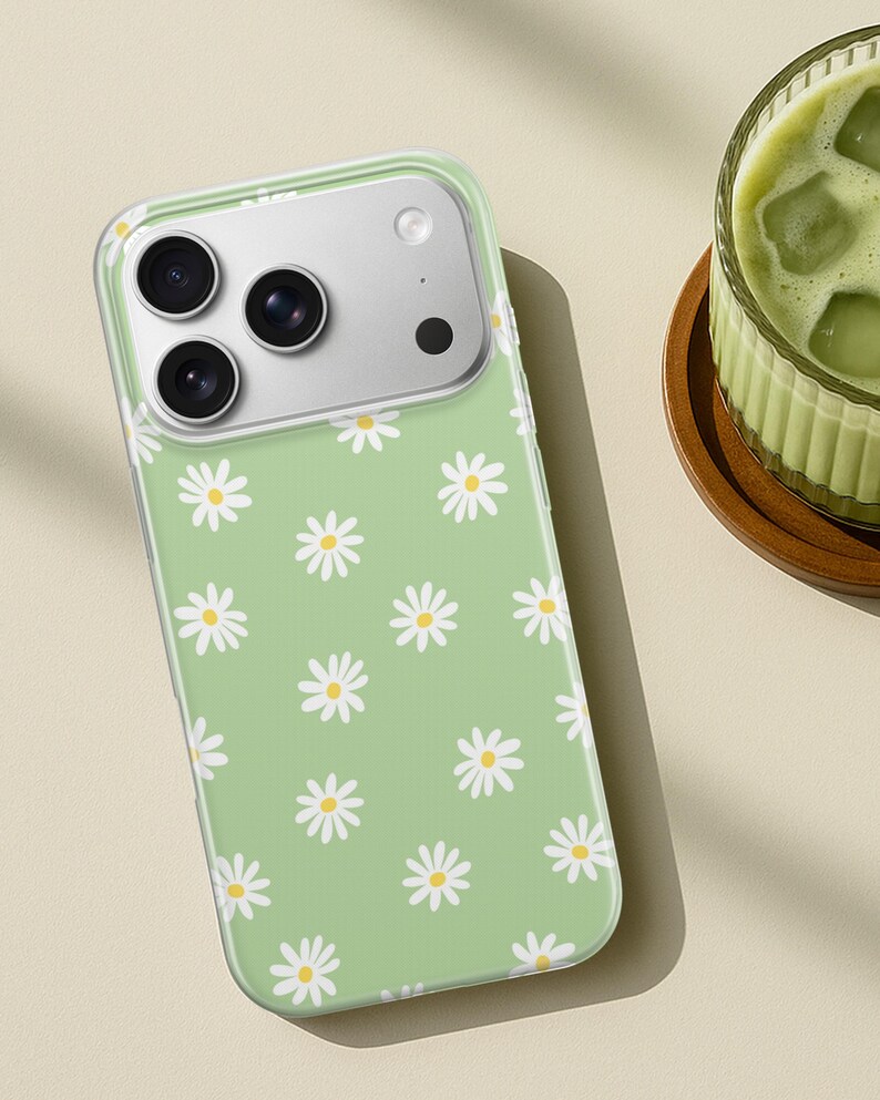Pode incluir: Uma capa de telefone verde claro com um padr&atilde;o de margaridas. A capa tem uma borda transparente e um recorte para a c&acirc;mera. As margaridas s&atilde;o brancas com centros amarelos. A capa est&aacute; sobre uma superf&iacute;cie marrom claro.