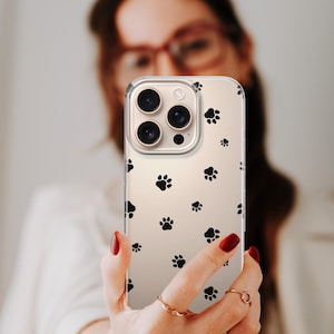 May include: Clear phone case with a pattern of black paw prints. The case is held up in front of a person. The phone case has a clear background and a protective design.