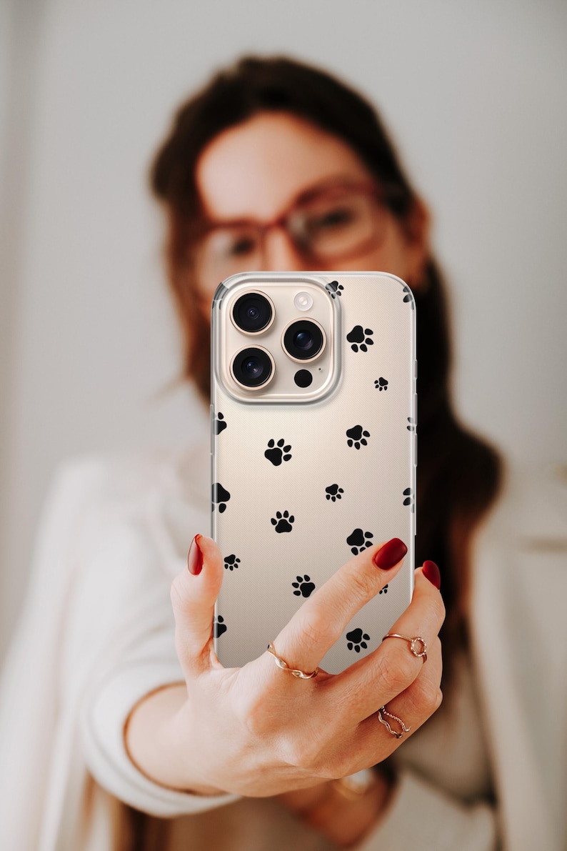 May include: Clear phone case with a pattern of black paw prints. The case is held up in front of a person. The phone case has a clear background and a protective design.