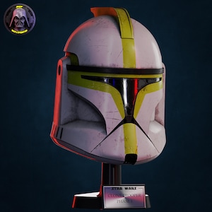 Puede incluir: Un casco de Clone Trooper Fase 1 blanco y amarillo de Star Wars, exhibido en un soporte negro. El casco tiene rayas amarillas y detalles en negro. La base tiene el logotipo de Star Wars y el texto "Clone Trooper Fase 1".