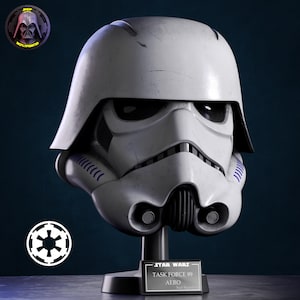 May include: A white Star Wars stormtrooper helmet with black accents and weathering marks. The helmet is mounted on a black stand with a plaque reading STAR WARS TASK FORCE 99 AERO. The design includes blue details on the sides and a black mouthpiece.