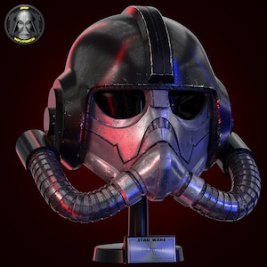 Op de afbeelding: Een zwarte en zilveren TIE-piloothelm met rood en blauw licht dat er vanaf reflecteert. De helm staat op een zwarte standaard met een plaquette waarop "STAR WARS TIE PILOT Ralph McQuarrie" staat.