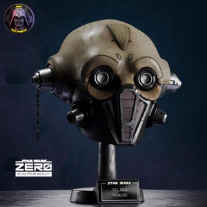 May include: A detailed replica of the Tel-Rea Vokoss helmet from Star Wars, featuring a tan dome, dark metallic accents, and circular eye lenses. The helmet is mounted on a black stand with a nameplate reading Star Wars Tel-Rea Vokoss.