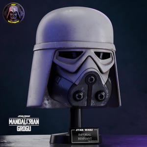 Puede incluir: Un casco gris Imperial Remnant de Star Wars: The Mandalorian and Grogu, exhibido en un soporte negro. El casco presenta un diseño detallado con una visera y un respirador. El logotipo y el texto de Star Wars son visibles.