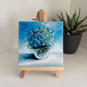 May include: A small, square painting on a wooden easel. The artwork depicts a bouquet of blue and yellow flowers in a clear glass vase against a light blue background. A potted succulent is in the background.