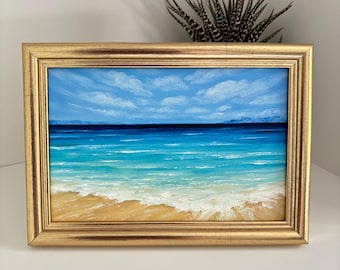 May include: A framed painting of a beach scene. The artwork depicts a bright blue sky with fluffy white clouds above a turquoise ocean. The waves crash on a sandy shore, all within a gold-colored frame.