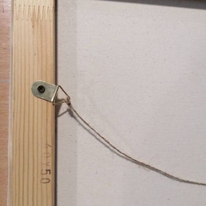 May include: A close-up of the back of a wooden framed canvas with a string attached to a metal hook. The canvas is white and has the dimensions 40x50 written on the frame.