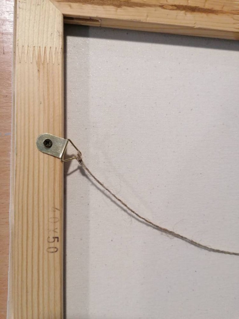 May include: A close-up of the back of a wooden framed canvas with a string attached to a metal hook. The canvas is white and has the dimensions 40x50 written on the frame.