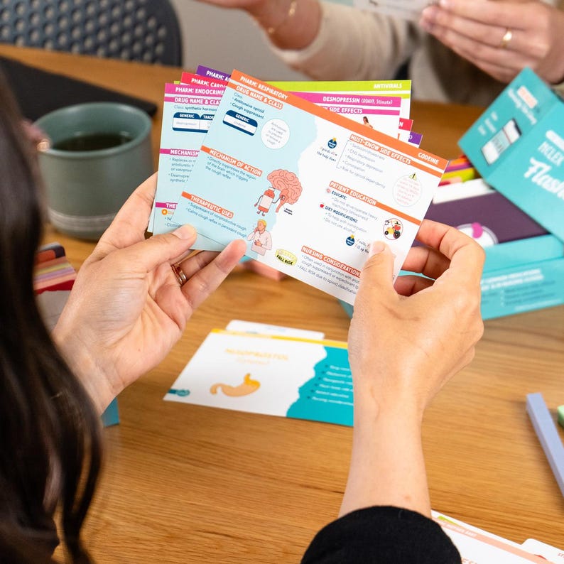 Puede incluir: Una persona sostiene una pila de tarjetas did&aacute;cticas coloridas con informaci&oacute;n m&eacute;dica. Las tarjetas presentan diagramas y texto sobre farmacolog&iacute;a, incluidos nombres de medicamentos, mecanismos de acci&oacute;n y usos terap&eacute;uticos. Una caja de tarjetas did&aacute;cticas es visible en el fondo.