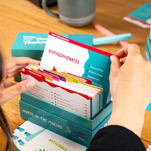 Puede incluir: Un juego de cajas de color turquesa de "Pharm Flashcards" con una pila de tarjetas. La tarjeta superior dice "Bisphosphonates" con un dise&ntilde;o rojo y blanco. La caja y las tarjetas est&aacute;n etiquetadas como "Nurse in the Making".