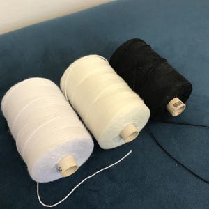 May include: Three spools of thread in white and black, on a dark blue surface. The spools are cylindrical, with thread wound around a cardboard core. The thread is unwound from the black spool.