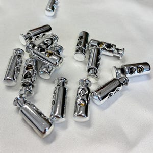 May include: A collection of silver-coloured cylindrical cord locks, each featuring a spring-loaded mechanism with two circular openings for threading cords. These metallic toggles have a smooth, reflective finish and are designed for securing drawstrings.