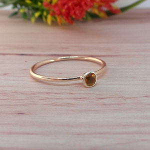 Può includere: Un delicato anello d'oro con una piccola gemma rotonda color ambra incastonata in una lunetta d'argento. L'anello è esposto su una superficie di legno chiaro, con una composizione floreale sfocata sullo sfondo.