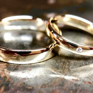 Puede incluir: Dos anillos de boda de oro. Uno es liso, mientras que el otro presenta un pequeño diamante redondo. Los anillos se asientan sobre una superficie metálica texturizada, creando un brillo cálido y reflectante. Las bandas son probablemente de oro amarillo.