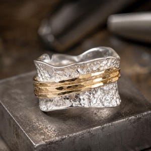 Puede incluir: Un anillo ancho de plata con una superficie martillada de textura pronunciada y bordes irregulares y ondulados. El centro de la banda está rodeado por dos hilos metálicos finos de color dorado pulido con textura martillada.
