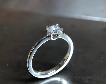 Könnte beinhalten: Ein silberner Verlobungsring mit einem runden, klaren Edelstein. Der Ring hat ein schmales Band und eine Vier-Krappen-Fassung, die den funkelnden Stein hervorhebt. Der Ring wird auf einer reflektierenden Oberfläche präsentiert.