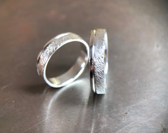 May include: Two silver rings with a textured, brushed finish. The rings have a smooth, polished interior and a band of texture around the exterior. The rings are likely wedding bands or promise rings.