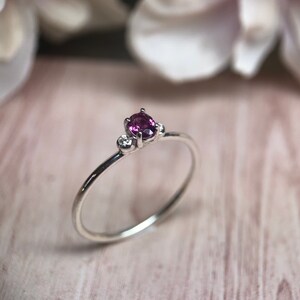 Könnte beinhalten: Ein silberner Ring mit einem zentralen, oval geschliffenen, tiefrosa Edelstein, flankiert von zwei kleineren, runden, klaren Edelsteinen. Der Ring hat einen dünnen Band. Der Hintergrund ist weich und verschwommen.