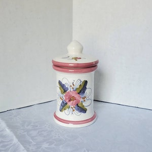 May include: White ceramic jar with a pink lid and floral design. The jar has a pink band around the top and bottom, and the floral design features a pink flower with blue and green leaves.