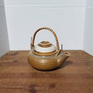 May include: A brown ceramic teapot with a white striped lid and a woven handle. The teapot has a brown glaze and a slightly textured surface.