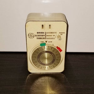 May include: A beige and brown GE Home Sentry Timer with a round dial that has numbers from 1 to 12. The dial has the words "NOON", "MORNING", "MIDNIGHT", and "EVENING" printed on it. The timer has three knobs: "AUTOMATIC", "MANUAL OFF", and "MANUAL ON".