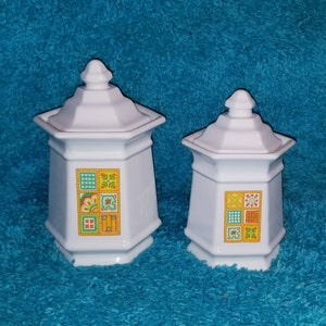 May include: Two white ceramic canisters with lids, featuring a colorful geometric pattern of squares and rectangles in shades of yellow, orange, green, and red.