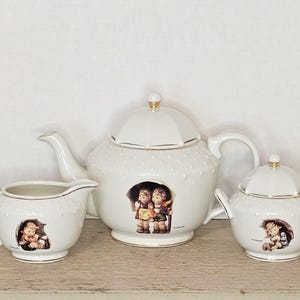 May include: A white porcelain tea set featuring a teapot, creamer, and sugar bowl. Each piece has a textured surface and is adorned with a colorful illustration of children under an umbrella. Gold trim accents the edges.