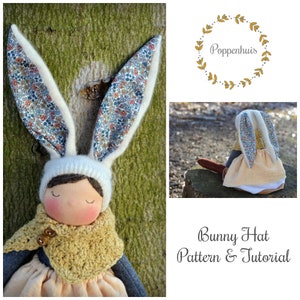 May include: A handmade doll wearing a white bunny hat with floral fabric ears and a crocheted scarf. The doll has brown hair, closed eyes, and a rosy complexion. The background is a blurred image of a tree trunk.