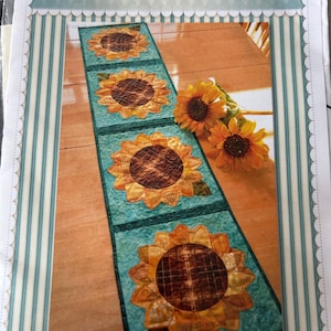 以下が含まれることがあります： 4つの大きなヒマワリのアップリケが付いた、パッチワークのヒマワリのテーブルランナー。ヒマワリは黄色の花びらと茶色の中心部があります。ランナーは、キルトのデザインのターコイズの背景にあります。「Shabby Fabrics」の文字が上部にあります。