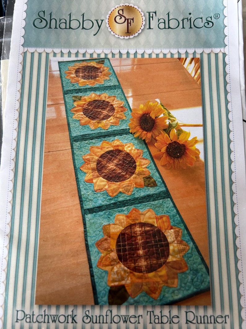 May include: A patchwork sunflower table runner featuring four sunflower appliques. The sunflowers have yellow petals and brown centres. The runner is on a teal background with a quilted design. The text "Shabby Fabrics" is at the top.