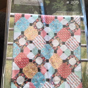 Op de afbeelding: Een quiltkit getiteld "Just Smashing #205" van Cotton Street Commons. De quilt heeft een kleurrijk patchwork-ontwerp met bloemen- en geometrische patronen in rood, blauw, geel en wit. De quilt wordt getoond op een metalen hek.