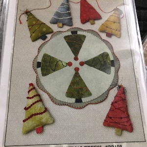 May include: A package featuring a Christmas-themed craft project. It includes several stuffed tree ornaments in various colors, such as green, red, and blue, with decorative stitching. The package also includes an 11-inch mat. The text on the package reads "Little Christmas Trees" and "5.5" Stuffed Tree Ornament or Garland Plus 11" Mat."