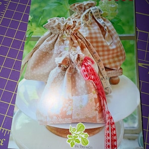 May include: A pattern booklet titled DITTY DRAWSTRING TRIO by Fig Tree & Co. featuring three fabric drawstring bags in beige, white, and orange floral and gingham prints. A beige measuring tape is draped over the bags, which are displayed on a white stand.